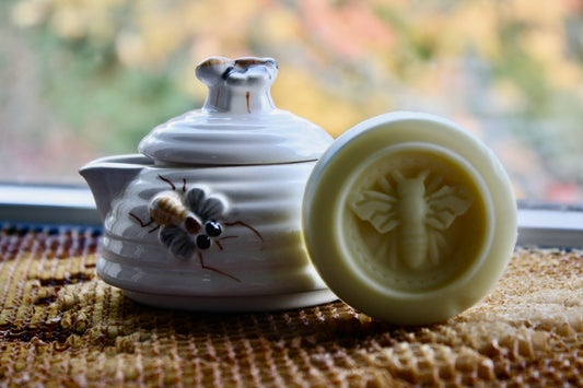 White ceramic teapot with bee design and a round white object with bee design on a textured surface.