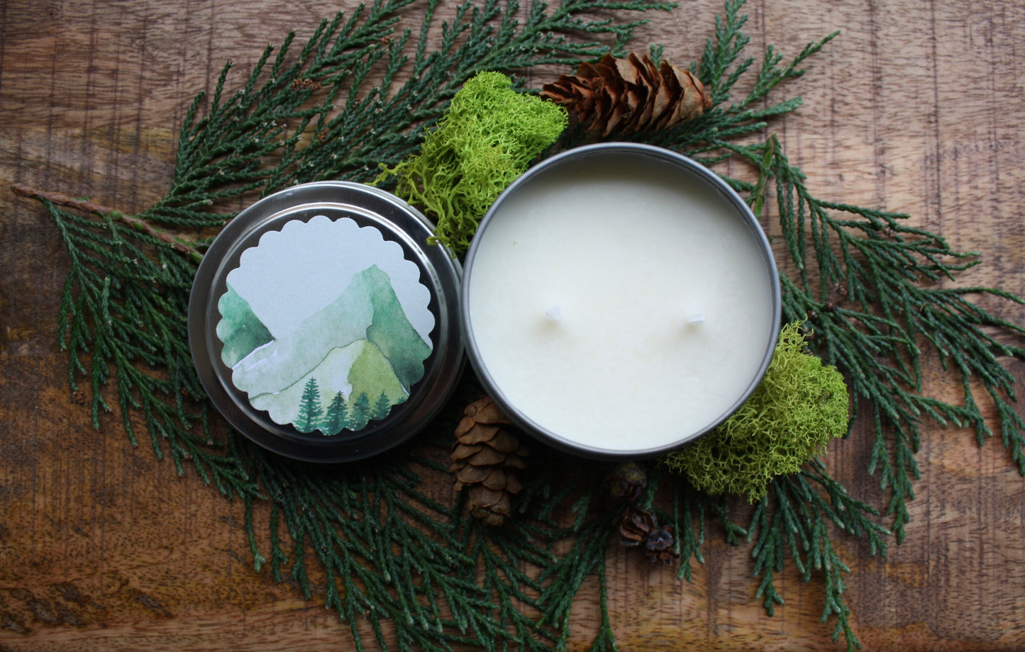 Candle in a tin with a decorative lid on a wooden surface with greenery