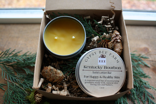 Box with a candle and a lotion bar labeled 'Kentucky Bourbon' on a wooden surface with greenery.