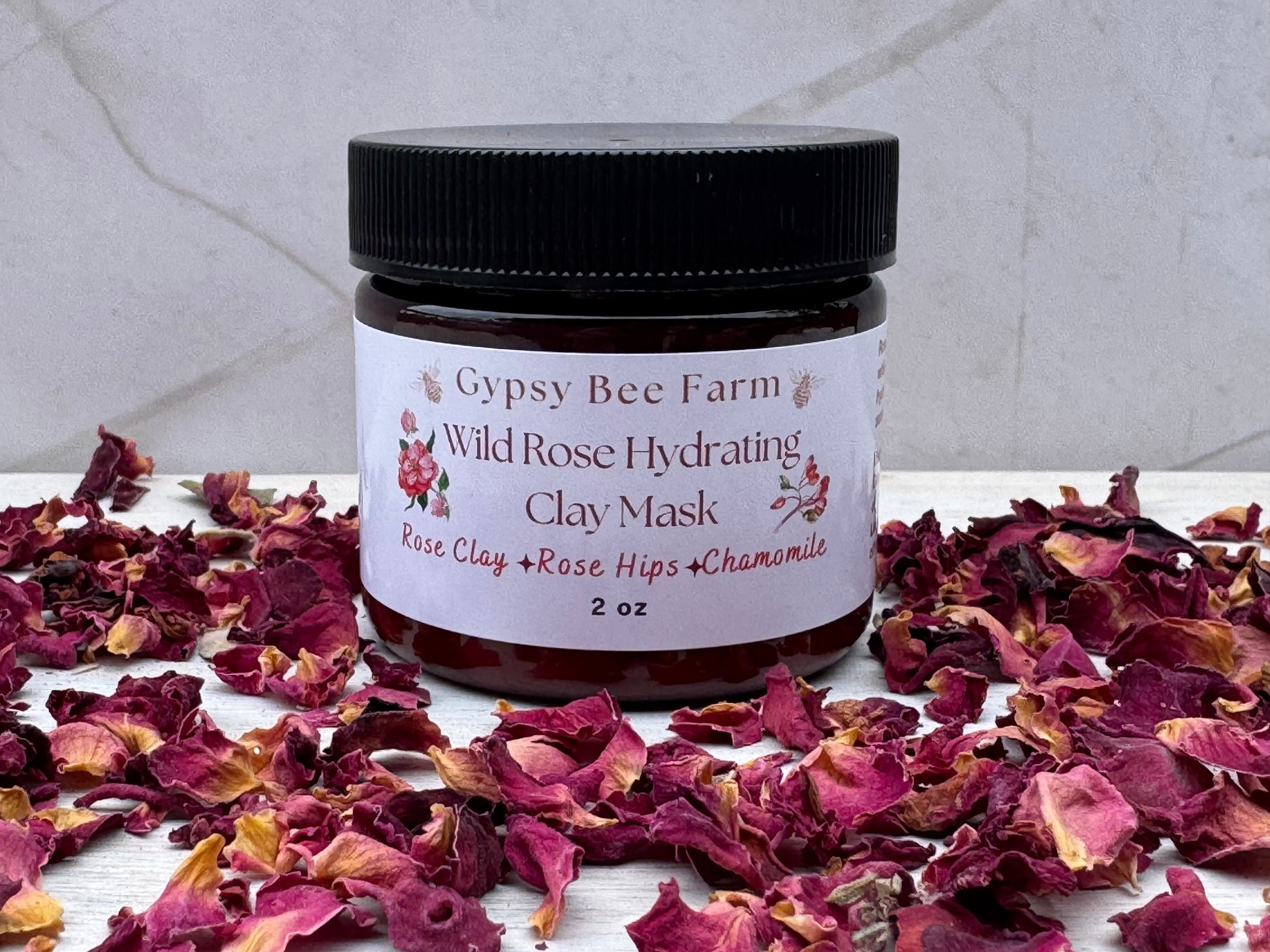 Gypsy Bee Farm Wild Rose Hydrating Clay Mask jar surrounded by dried rose petals on a white surface with a gray marble background.